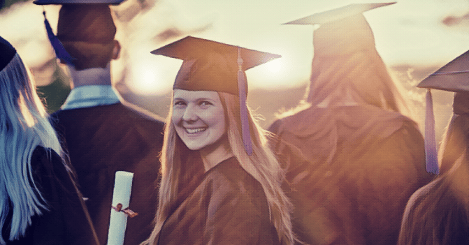 a graduating senior in full regalia clutches her diploma and smiles broadly as she turns to look at the camera while she and her classmates stand as a group, they with their backs to the camera.