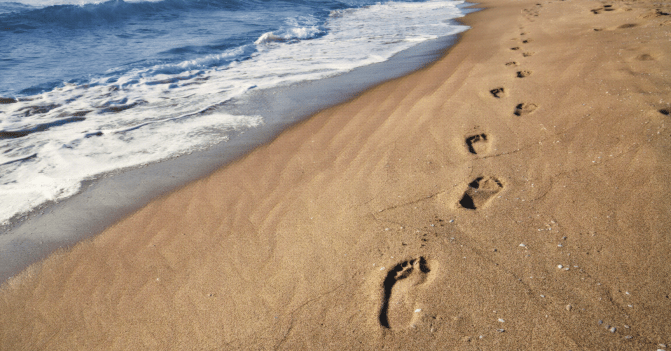 A beach scene in which footprints line the edge of the water, destined to disappear with the incoming waves and vanish without a trace