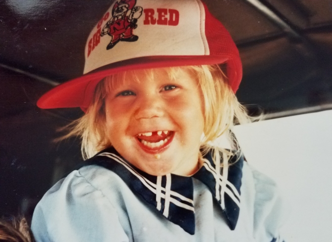 A little girl with blond hair, missing a front tooth, and wearing a Nebraska Cornhuskers baseball cap too big for her