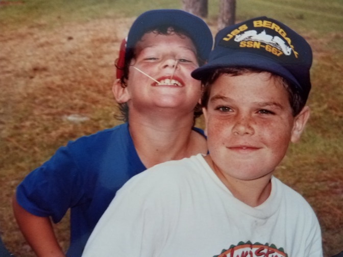 Two boys wearing baseball caps. One has toothpicks sticking out of his big grin. The other is sporting his father's submarine squadron cap.