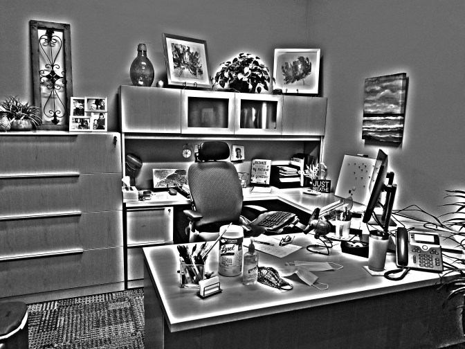 an office at a workplace, not an office at home. The desk bears Lysol wipes, hand sanitizer, and face masks, in addition to the usual work supplies