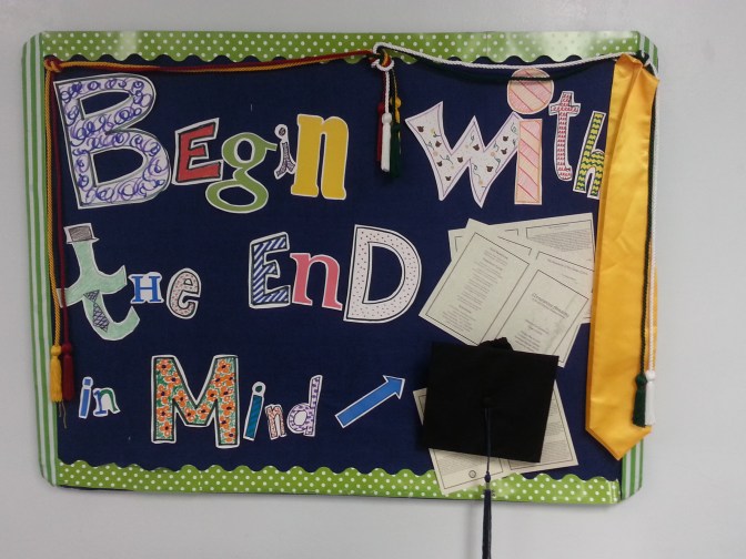 A bulletin board with the message "Begin with the end in mind," complete with a graduation cap and program, honors cords and scarf