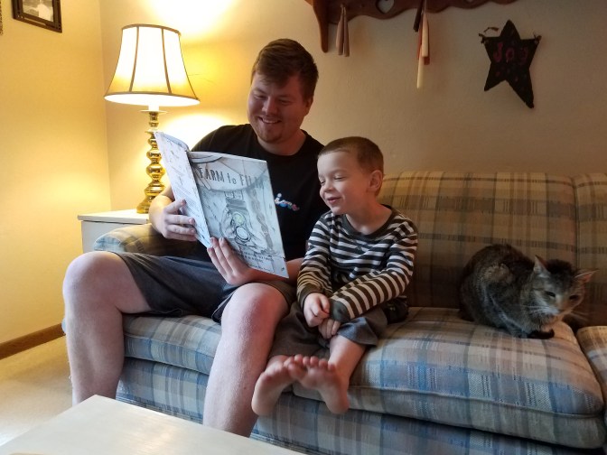 An uncle reads Farm to Film to his young nephew. A cat sits beside them on the couch.