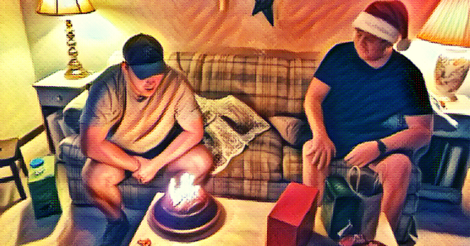 My oldest son blows out the candles on a birthday cake baked by my youngest son, sitting at his right with a Santa hat on his head.