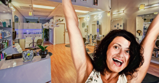a woman with brown eyes and brown curly hair jumps for joy in front of a hair salon background.