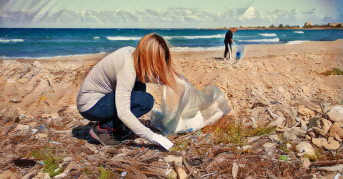a woman in the foreground of this seaside image, wears gloves as she picks up debris and puts it in a trash bag. A man in the distance is walking with a trash bag, looking down at the beach sand for trash.
